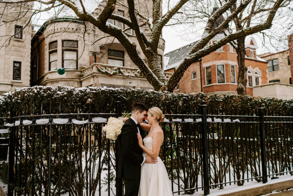 Bride and groom stand on a sidewalk in front of Stan Mansion in Chicago in the winter. There is snow on the ground and the bushes behind them. 