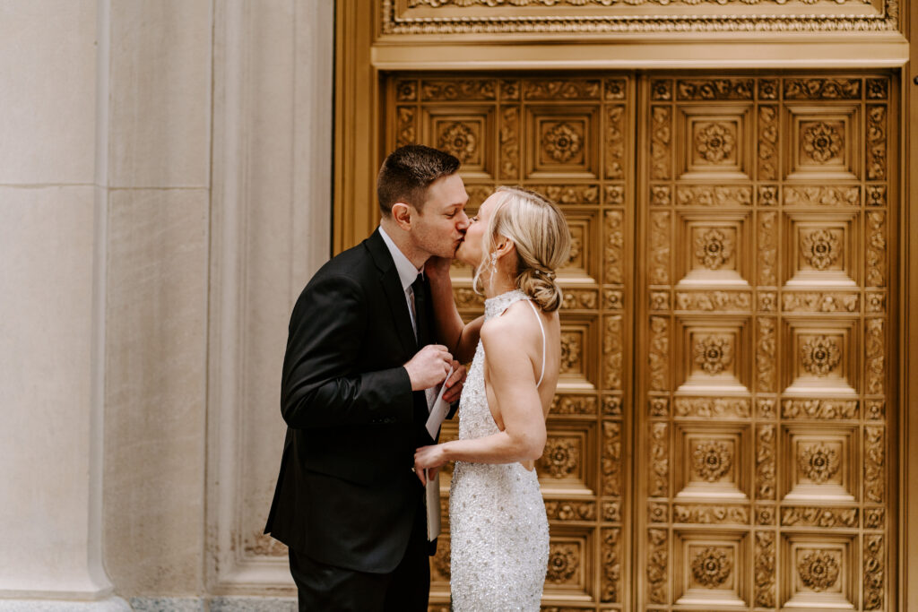 Bride in a glittery white halter dress and groom in a black suit kiss in front of golden ornate doors to the Federal Reserve Building during their intimate vows at their elopement ceremony in Chicago. 