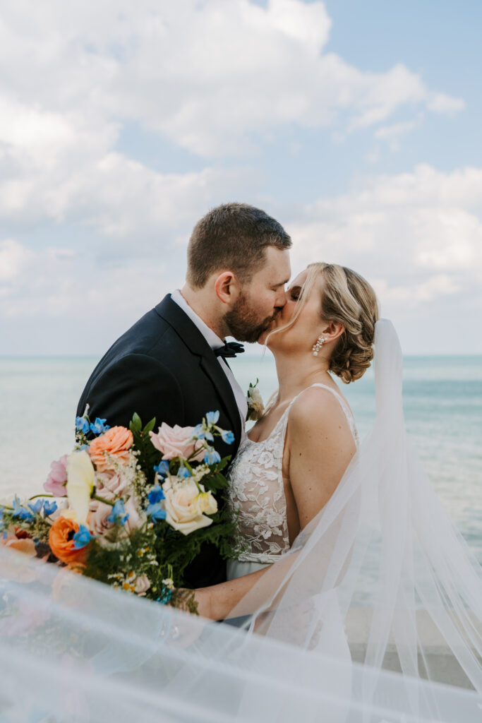 Bride and groom kiss as the bride holds a colorful bouquet of yellow, orange and blue flowers. They stand at the Chicago Lakefront with Lake Michigan in the background. 