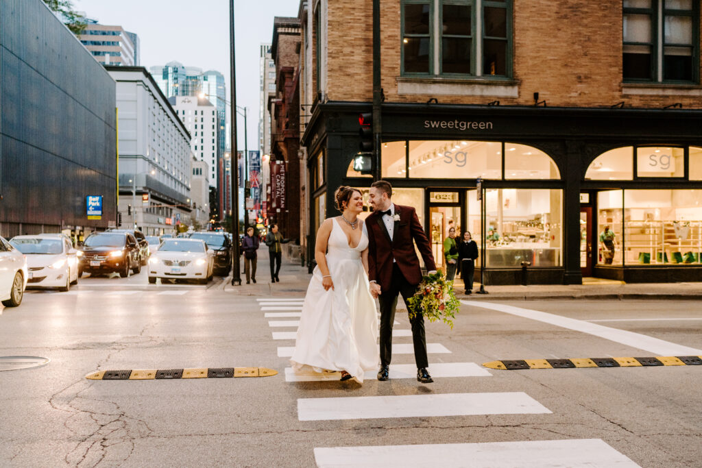 Groom in a red suit jacket and bow tie leans into his bride in a deep cut white dress as they cross the street in downtown Chicago. Sweetgreen is in the background of the street as cars drive beside them. 