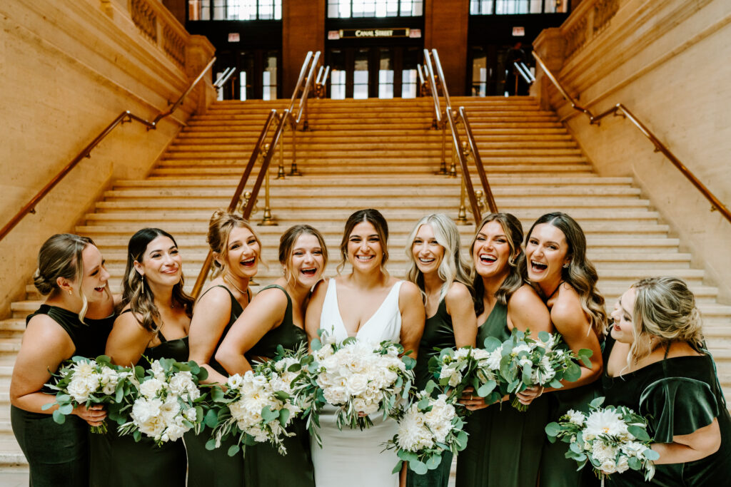 Bride smiles as her bridesmaids in green dresses lean in and laugh candidly. They are standing at the bottom steps of Union Station in Chicago. 