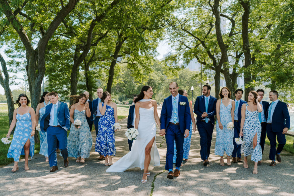 Bride in a white strapless dress and a scarf walks look at her groom in a blue suit. Her bridesmaids and his groomsmen walk behind them candidly looking at each other and smiling. They are walking through Lincoln Park in Chicago.