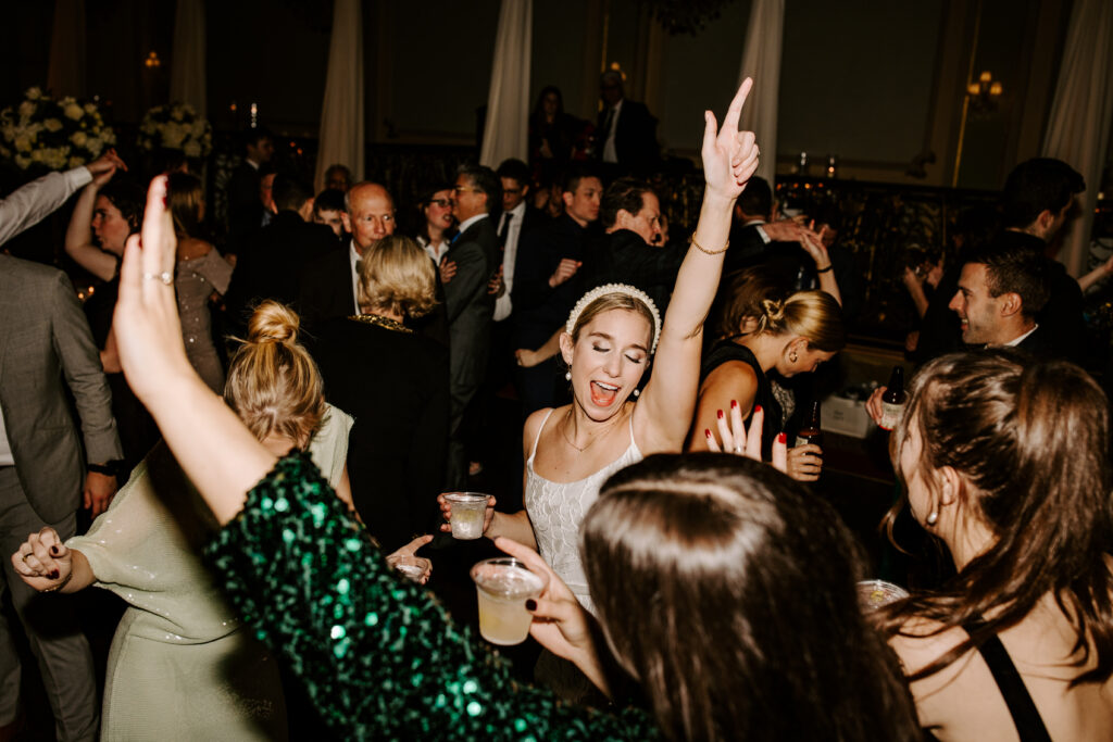 Bride in a pearl head band sings and places one arm in the air as she dances during the reception surrounded by other dancing guests during her Chicago Wedding Reception. 