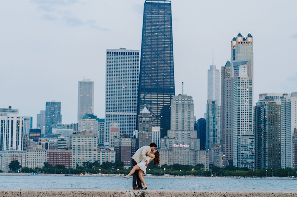 A couple kisses and dips during their engagement session on the ribbon at North Avenue Beach, one of the best places to see the Chicago skyline along Lake Michigan.