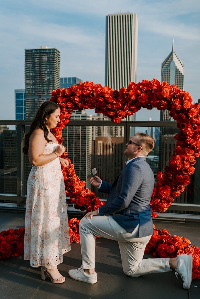 A groom proposes to his bride on a rooftop in Chicago with a heart made out of roses behind him.  The fiancée wears a light pink and blue textured cotton dress and he wears khaki pants and a blue sport coat.