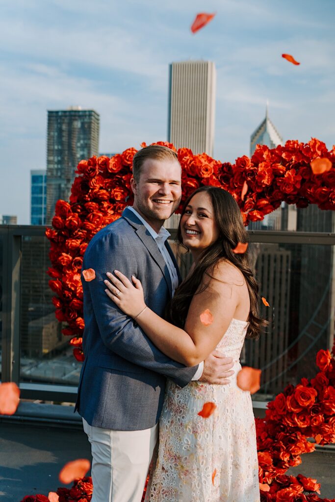 A groom proposes to his bride on a rooftop in Chicago with a heart made out of roses behind him.  The fiancée wears a light pink and blue textured cotton dress and he wears khaki pants and a blue sport coat.  The Chicago wedding photographer throws roses around them to celebrate.