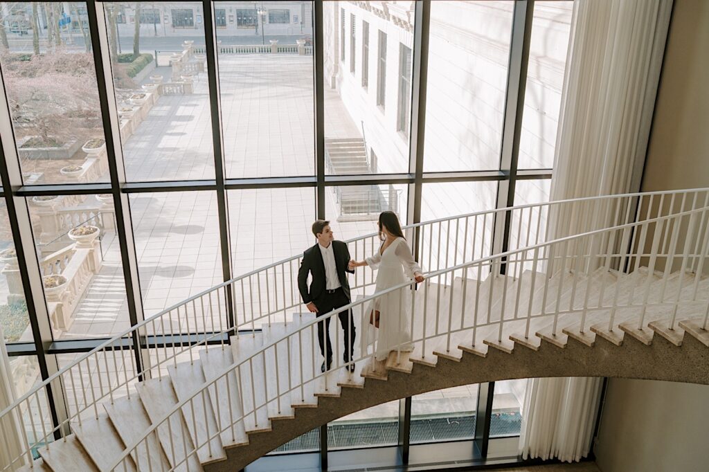 A couple walks down the stairs overlooking the back patio at the Art Institute in Chicago.  The fiancée wears a white sheer dress and holds her fiancé's hand as they walk down the marble staircase.