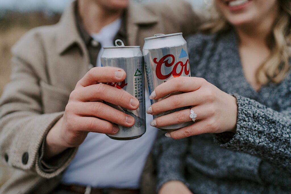 A fiancée's toast their engagement during their engagement session in the Lincoln Park Boardwalks with Coors Lights.  You can see the brides oval cut diamond engagement ring with pear stones on either side of it.