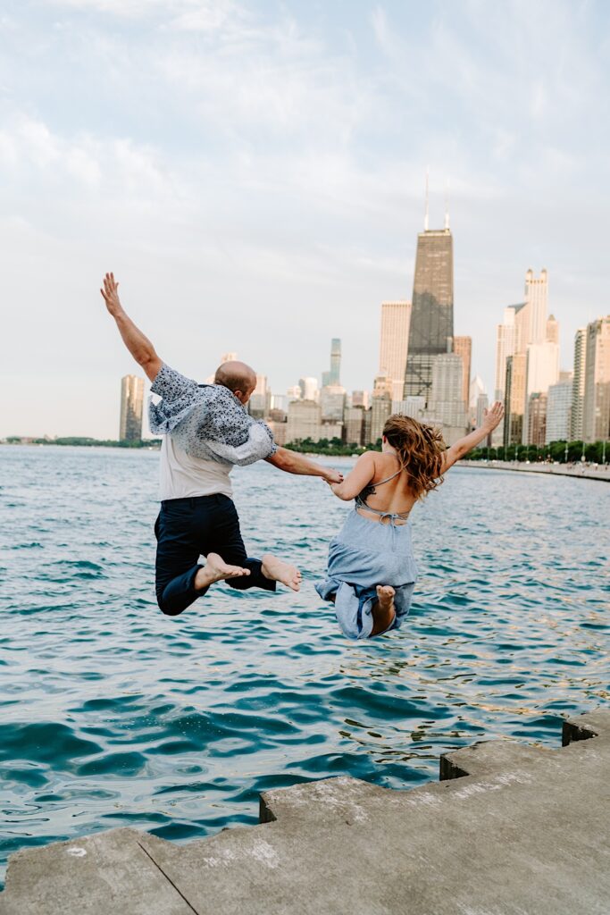 A couple jumps into the water of Lake Michigan overlooking the skyline of Chicago at North Avenue Beach.  They are both wearing blue matching the Chicago summer sky.