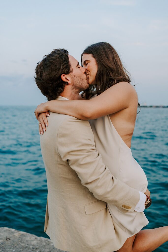 A couple wearing beige outfits, kisses while standing  on the ribbon overlooking Lake Michigan at North Avenue Beach in Chicago. 