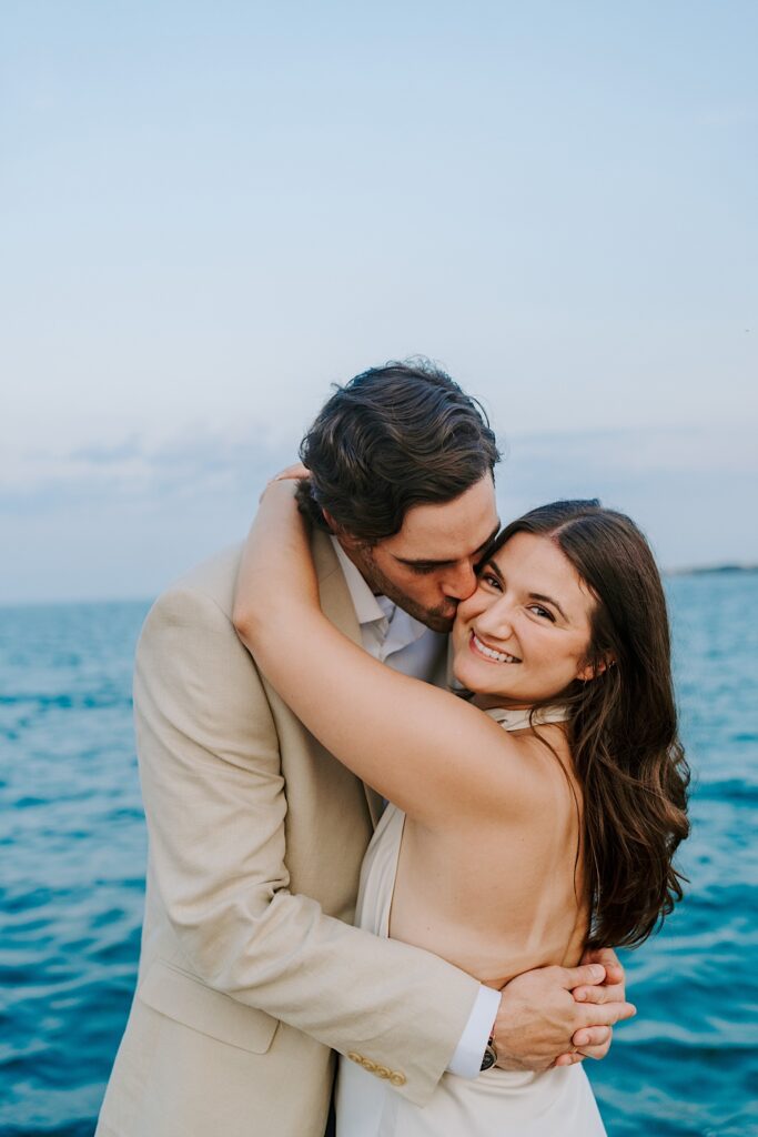 A fiancé kisses his soon to be wife on the cheek while she looks towards her Chicago wedding photographer.  They are taking engagement photos at North Avenue Beach during the summer.