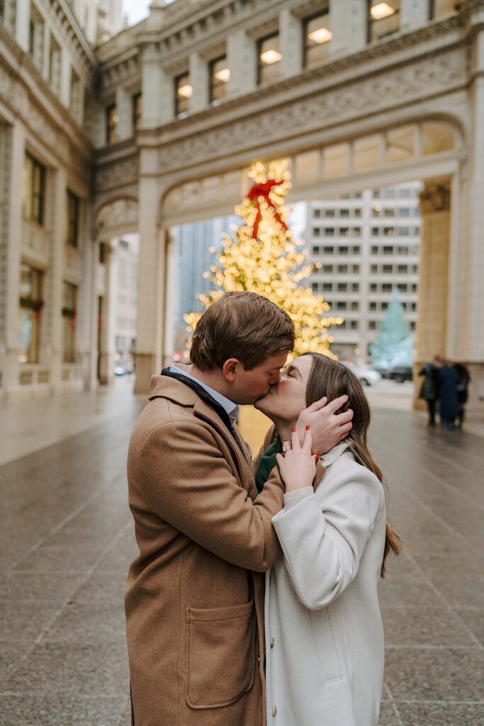 A couple kisses during their engagement session during the winter in Chicago.  In the background you can see a lit up Christmas tree with a red ribbon on top underneath the Wrigley Building in Chicago. 