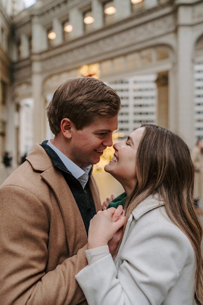 A couple stands smiling at eachother during their engagement session during the winter in Chicago.  In the background you can see a lit up Christmas tree with a red ribbon on top underneath the Wrigley Building in Chicago. 