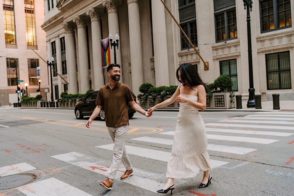 A couple in chic neutral colored outfits walk across the street during their engagement session down the street from the Chicago Board of Trade Building. 