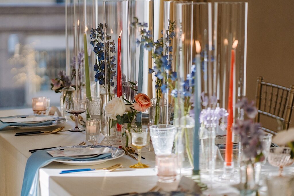 A table of a wedding reception decorated with tall colorful candles, tableware, and colorful florals